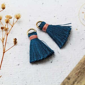 Op de afbeelding: Twee teal blauwe kwastcharms met gouden springringen en een roze accent. De kwasten bevinden zich op een witte achtergrond.