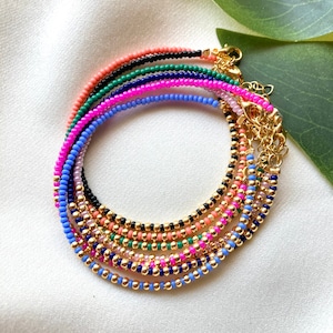 May include: A set of colorful beaded bracelets with gold accents. The bracelets are layered and feature different colors, including pink, blue, green, and black.