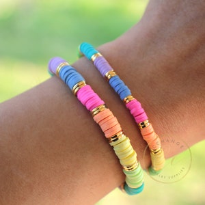 May include: Two colourful bracelets with gold accents. The bracelets are made of small, round beads in shades of blue, purple, pink, orange, yellow, and green. The bracelets are stacked on a person's wrist.