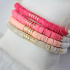 May include: Stack of four bracelets with pink, light pink, and white ceramic beads. The beads are separated by gold spacers. The bracelets are on a white surface.