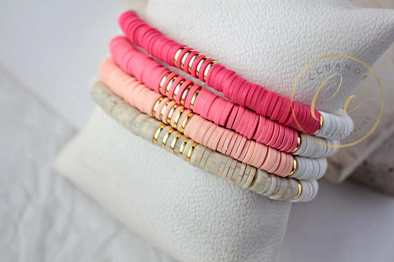 May include: Stack of four bracelets with pink, light pink, and white ceramic beads. The beads are separated by gold spacers. The bracelets are on a white surface.