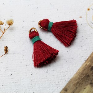 Op de afbeelding: Twee rode kwastbedels met gouden springringen en groene trim. De kwasten zijn gemaakt van zacht, pluizig garen en zijn perfect om een ​​toets van whimsy toe te voegen aan sieraden of knutselwerk.