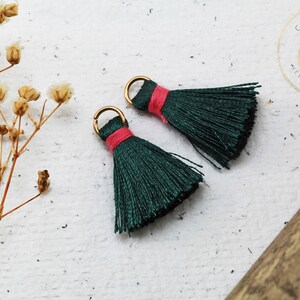 Op de afbeelding: Twee groene kwastcharms met roze trim en gouden springringen. De kwasten zijn gemaakt van zacht garen en zijn ongeveer 2,5 cm lang.