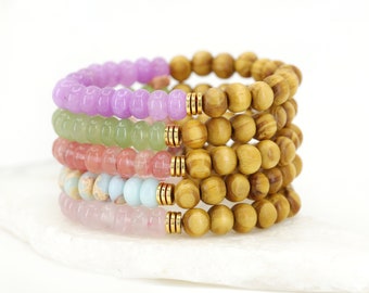 May include: A stack of colorful beaded bracelets, including purple, green, pink, and blue, with gold accents. The bracelets are made of wood and gemstone beads.