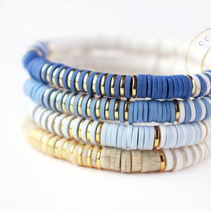 May include: Four stacked bracelets with blue, white, and gold beads. The bracelets are made of ceramic and have a layered ombre effect.