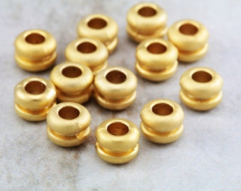 Könnte beinhalten: Eine Gruppe von 12 goldfarbenen Metallperlen, jede mit einem runden Loch in der Mitte. Die Perlen sind in einem zufälligen Muster auf einer hellgrauen Oberfläche angeordnet.