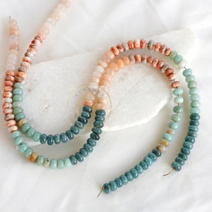 May include: Two strands of gemstone beads in shades of peach, orange, green and teal. The beads are strung on a thin wire. The beads are displayed on a white surface. The beads are measured in centimetres.