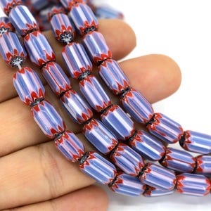 Può includere: Fili di perline di vetro a righe blu e bianche con accenti rossi. Le perline sono cilindriche con un motivo a spina di pesce e sono infilate insieme. Le perline sono tenute in una mano su sfondo bianco.