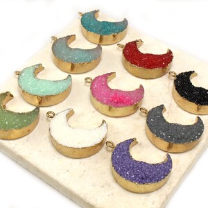 May include: Assortment of ten gold-rimmed crescent moon shaped pendants with various colored crystal accents. The pendants are arranged in a row on a white surface.