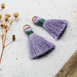 Op de afbeelding: Twee lavendelkleurige kwastjes met gouden ringen en groene accenten. De kwastjes liggen op een wit oppervlak.