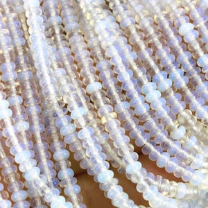 May include: Multiple strands of small, translucent, round beads with a pale blue and opalescent sheen. The beads are strung on thin, clear filaments, creating a dense, uniform texture of light-reflecting, smooth, circular shapes arranged in parallel lines.