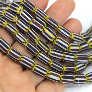 Puede incluir: Primer plano de hilos de cuentas de colores. Las cuentas tienen un patrón repetitivo de rayas marrones, blancas y negras, con detalles amarillos. Las cuentas cilíndricas son probablemente de vidrio o un material similar. Las cuentas se sostienen en una mano.