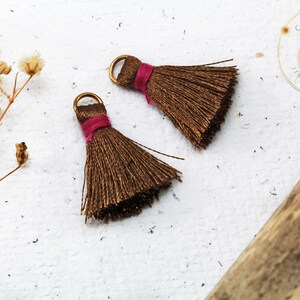 Op de afbeelding: Twee bruine kwastjes met gouden metalen ringen en roze draadaccenten. De kwastjes liggen op een wit oppervlak.