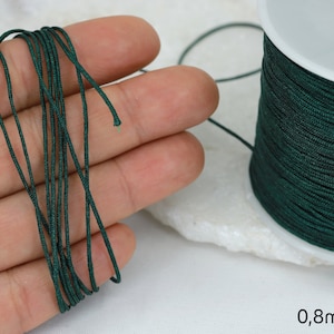 Puede incluir: Una bobina de cordón elástico verde oscuro de 0,8 mm de grosor, con una mano sujetando un trozo de cordón.
