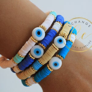 Può includere: Un set di quattro braccialetti con perline in ceramica blu, bianca e beige. Ogni braccialetto è decorato con un ciondolo a forma di occhio blu con un bordo dorato.