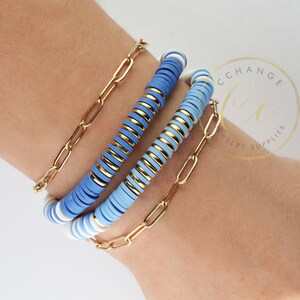 May include: A set of three bracelets featuring blue and white beads with gold accents. The bracelets are stacked on a person's wrist.