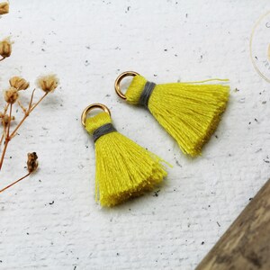 Op de afbeelding: Twee gele kwastcharms met gouden springringen en grijze trim. De kwasten zijn ongeveer 5 cm lang.