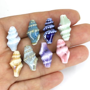 May include: A collection of nine ceramic seashell beads in various colors, including blue, green, pink, and yellow. The beads are held in a hand, showcasing their detailed spiral designs and glossy finish. These beads are ideal for jewelry making.