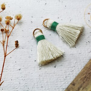 Op de afbeelding: Twee witte kwastbedels met groene bies en gouden springringen. De kwasten zijn gemaakt van zacht, pluizig garen en zijn perfect om een ​​toets van whimsy toe te voegen aan sieraden of knutselwerk.