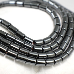 Peut inclure: Gros plan de plusieurs brins de perles cylindriques gris foncé. Les perles sont enfilées ensemble, formant une forme incurvée sur un fond blanc. Les perles ont un éclat métallique et une texture lisse, suggérant qu'elles sont faites d'un matériau poli.