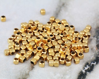 May include: A pile of gold-colored metal beads, each bead is a cube with rounded edges. The beads are scattered on a white marble surface.