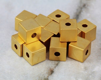May include: A group of gold-colored metal cube beads with a hole in the center of each cube. The beads are arranged in a pile on a marble surface.