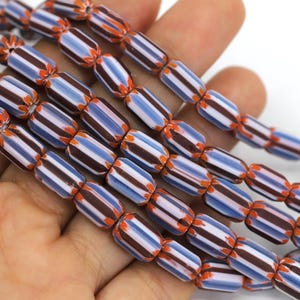 Puede incluir: Primer plano de hilos de cuentas a rayas. Las cuentas cilíndricas presentan rayas blancas, marrones y azul claro, con detalles en naranja. Las cuentas se sostienen en una mano sobre un fondo blanco.
