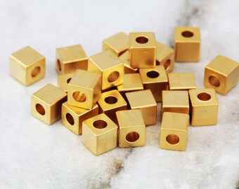 May include: A pile of gold-colored metal cube beads with a hole in the center of each cube. The beads are scattered on a white surface.