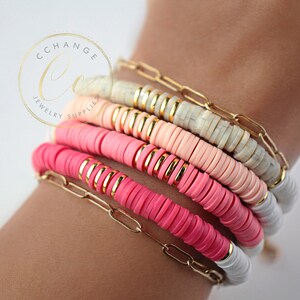 May include: A stack of colorful bracelets with pink, white, and peach beads. The bracelets are made of round beads with gold accents and are layered on a person's wrist.