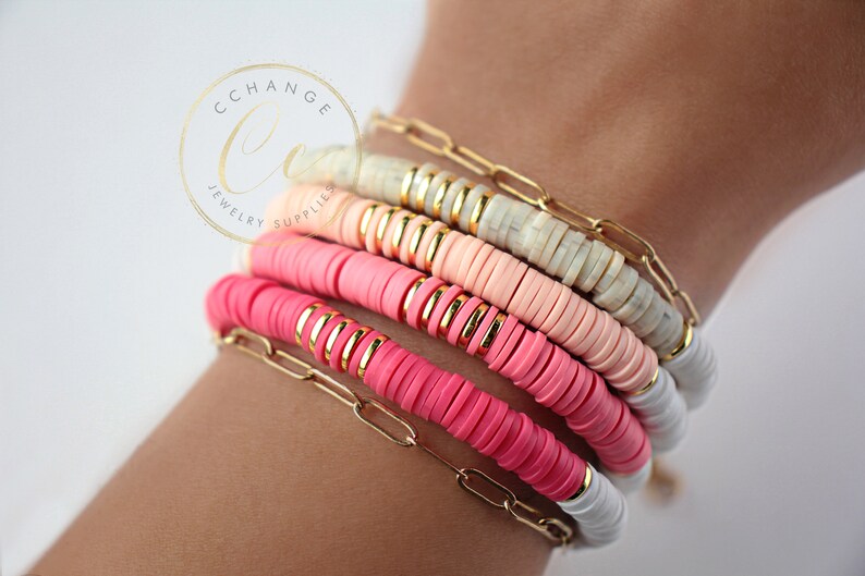 May include: A stack of colorful bracelets with pink, white, and peach beads. The bracelets are made of round beads with gold accents and are layered on a person's wrist.