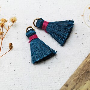 Op de afbeelding: Twee teal blauwe kwastcharms met gouden springringen en roze draadaccenten. De kwasten liggen op een wit oppervlak.