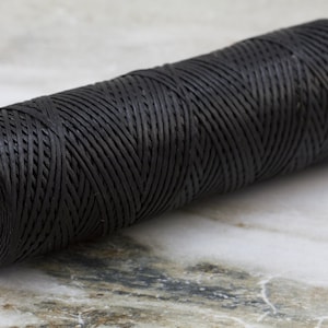 Może przedstawiać: Szpula czarnej, woskowanej nici do obróbki skóry. Nić jest ciasno nawinięta na czerwoną plastikową szpulę.