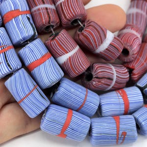 Pode incluir: Close-up de uma coleção de contas de vidro coloridas. As contas são cilíndricas com um padrão listrado em azul, vermelho e branco. Cada conta é acentuada com uma faixa vermelha. As contas são enfiadas, mostrando uma paleta de cores vibrantes.