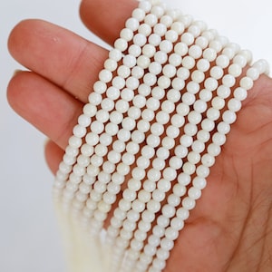 May include: A strand of white round beads made from natural mother of pearl. The beads are smooth and have a subtle iridescent sheen.