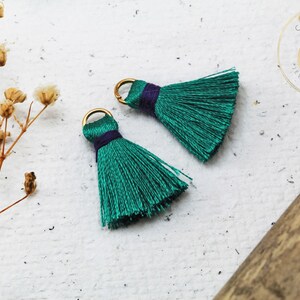 Op de afbeelding: Twee teal groene kwastversieringen met gouden springringen en paarse accenten. De kwasten zijn gemaakt van zachte, zijdeachtige draden en zijn perfect om een ​​toets van elegantie aan elk project toe te voegen.
