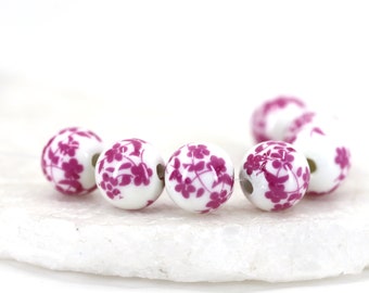 May include: A group of six white ceramic beads with a pink floral design. The beads are round and have a hole in the center for stringing.