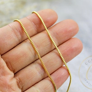 May include: A gold chain necklace with a box chain design. The chain is shown on a hand, with the ends of the chain hanging down.