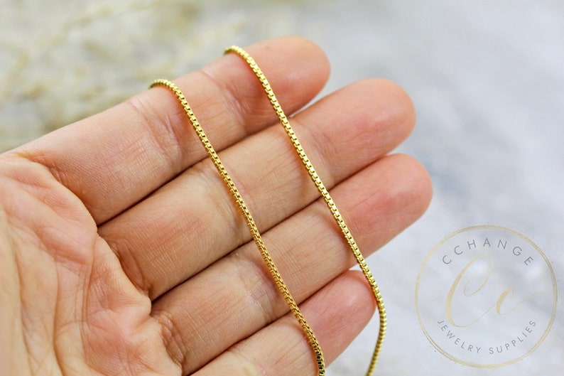 May include: A gold chain necklace with a box chain design. The chain is shown on a hand, with the ends of the chain hanging down.