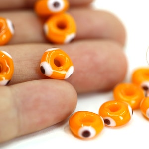Puede incluir: Cuentas de vidrio naranja con un diseño de ojo turco blanco y negro. Las cuentas tienen un agujero en el centro para enhebrar.