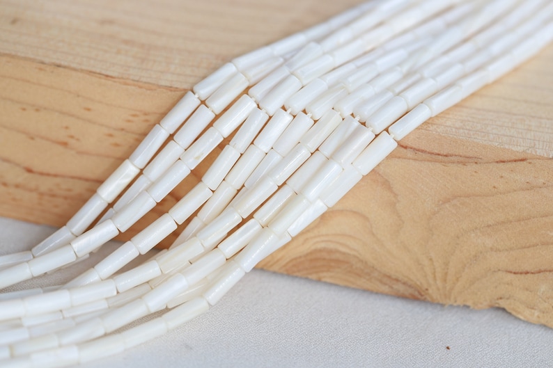 Pu&ograve; includere: Una collana di perline bianche di conchiglia, ogni perlina ha una forma cilindrica. Le perline sono disposte in fila su una superficie di legno marrone chiaro.