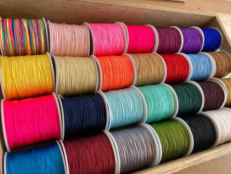 May include: A collection of spools of colourful waxed cord, arranged in a wooden box. The cords are in various colours, including red, blue, green, yellow, pink, and purple.