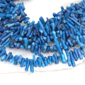 May include: A strand of blue coral beads, each bead is a long, thin, and slightly curved piece of coral. The beads are arranged in a loose, flowing pattern.