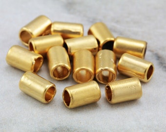 Könnte beinhalten: Eine Gruppe von 14 goldfarbenen Metallperlen, jede mit einer zylindrischen Form und einer glatten, glänzenden Oberfläche. Die Perlen sind in einem lockeren Haufen auf einer Marmorfläche angeordnet.