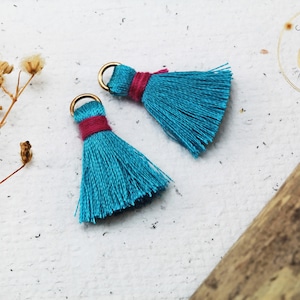 Op de afbeelding: Twee turquoise blauwe kwastbedels met gouden springringen en een roze accent. De kwasten zijn gemaakt van zacht, pluizig garen en zijn perfect om een vleugje whimsy toe te voegen aan sieraden of knutselwerk.