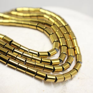 Può includere: Un filo di perline di ematite placcate oro, ogni perlina è un cilindro con un bordo leggermente arrotondato. Le perline sono disposte in un motivo circolare, creando un aspetto simile a una collana.