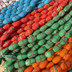 Pode incluir: Múltiplos fios de contas de formato oval dispostos em fileiras. As contas apresentam um padrão de cruz em cores contrastantes, incluindo branco, azul claro, vermelho, verde e laranja. Cada conta tem um acabamento liso e brilhante.