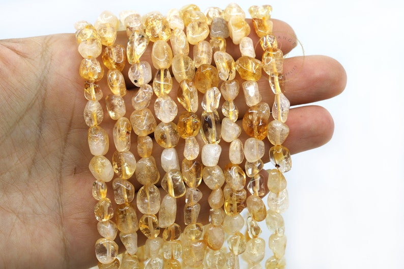 May include: A strand of tumbled citrine gemstone beads in various shades of yellow and orange. The beads are smooth and polished, with a natural, irregular shape.