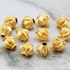 May include: A group of 12 gold-coloured metal beads with a textured, fluted design. The beads are arranged in a circle on a white marble surface.