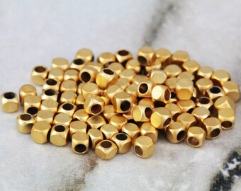 May include: A pile of gold-colored metal beads with a hexagonal shape and a hole in the center. The beads are scattered on a light gray marble surface.