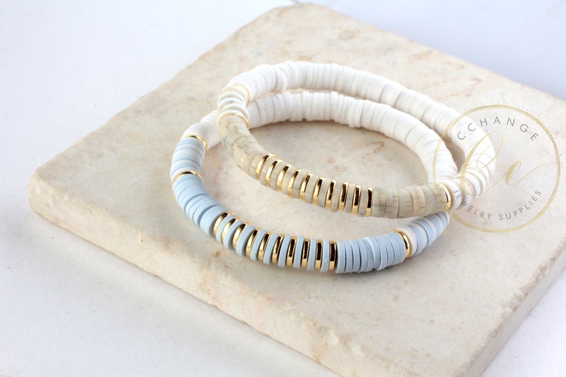 May include: Two bracelets made of white and light blue ceramic beads with gold accents. The bracelets are stacked on top of each other.